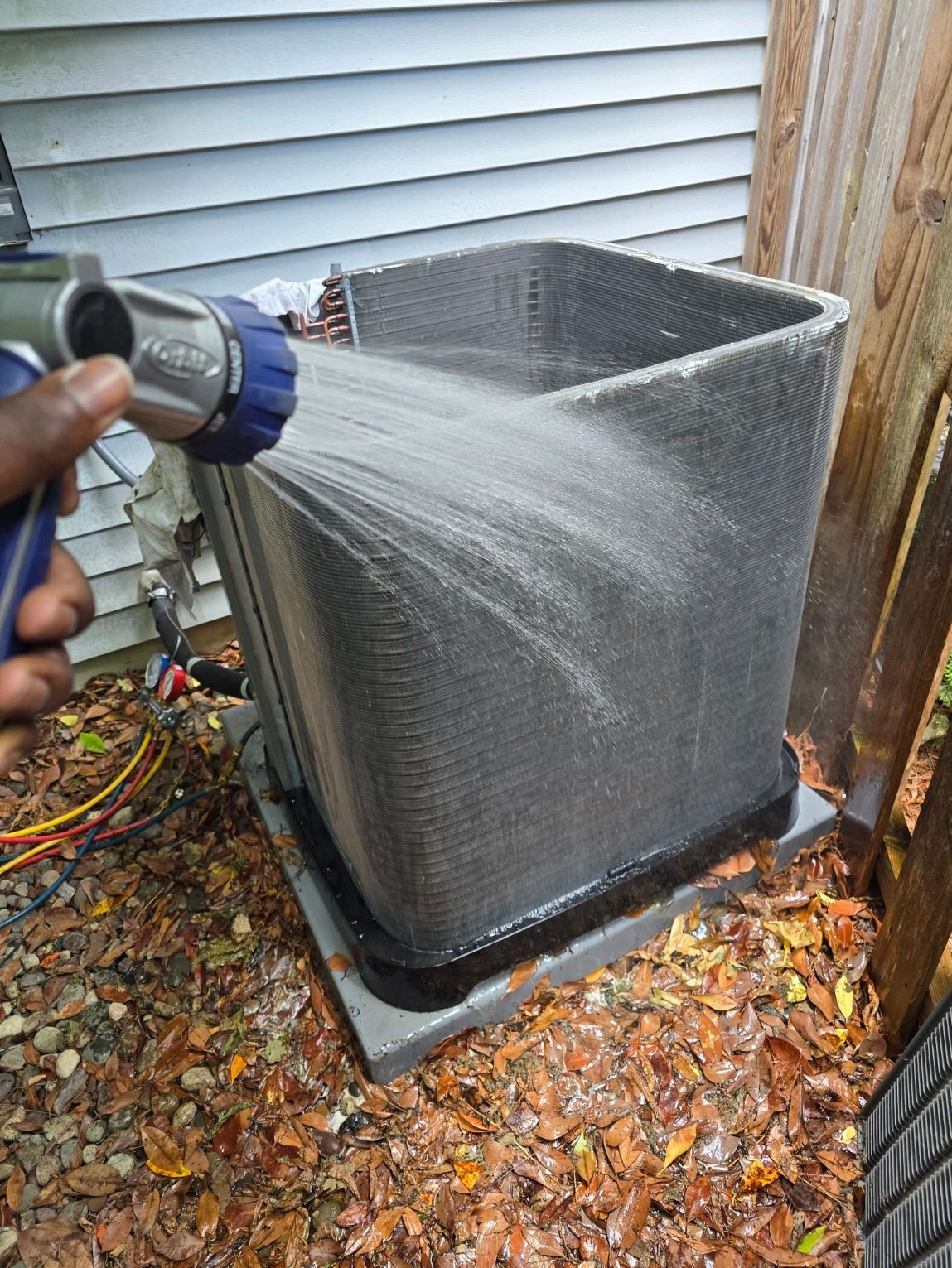 Person using a spray nozzle to clean an outdoor air conditioning unit surrounded by fallen leaves.