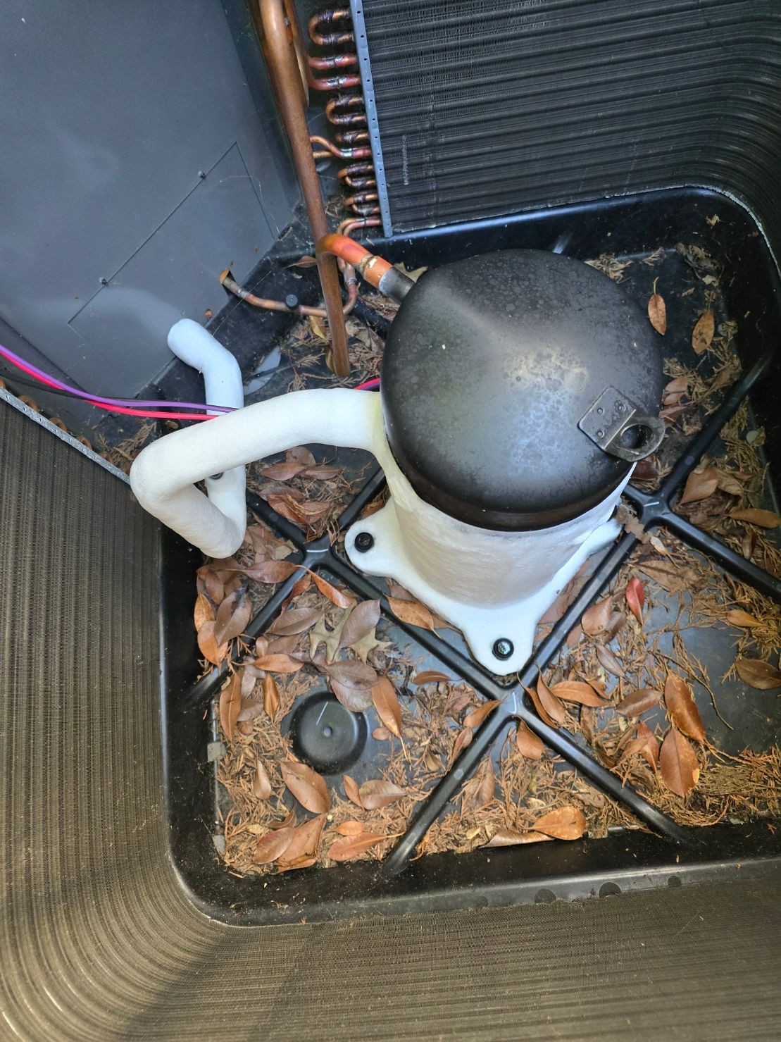 Close-up of an air conditioning unit compressor with frost on the pipes and surrounded by fallen leaves.