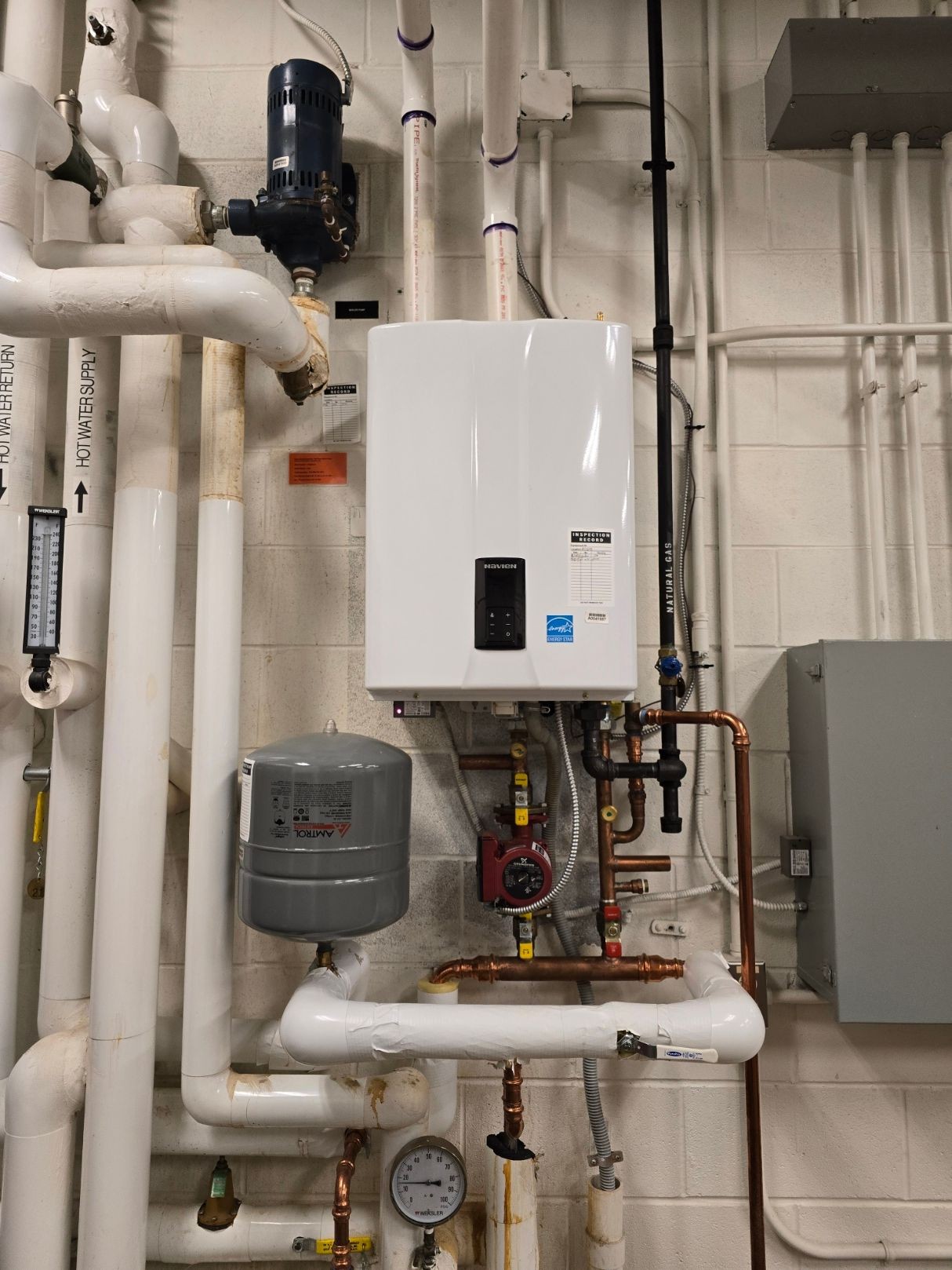 Industrial boiler system with various pipes, valves, and gauges installed on a white brick wall.